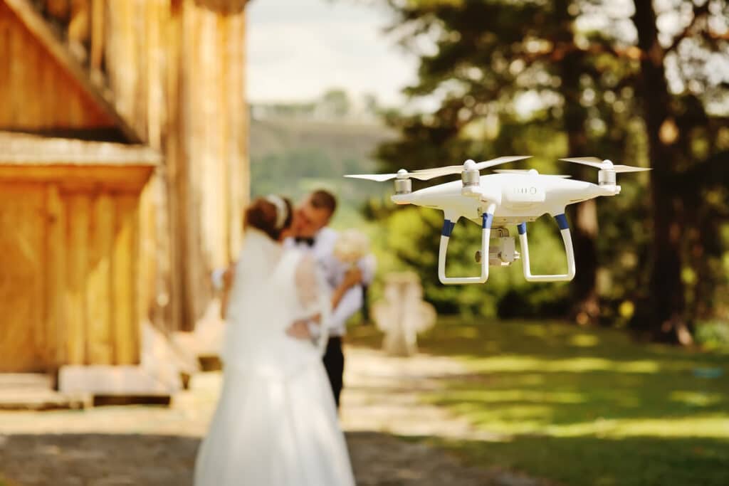 Drone mariage : Capturez des moments inoubliables Drone blanc filme un couple de mariés s'embrassant en arrière-plan flou, dans un cadre extérieur verdoyant près d'un bâtiment en bois.
