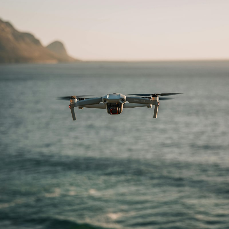 Drone gris DJI Mavic Air 2 volant au-dessus de l'océan calme au lever/coucher du soleil, montagnes floues à l'arrière-plan.