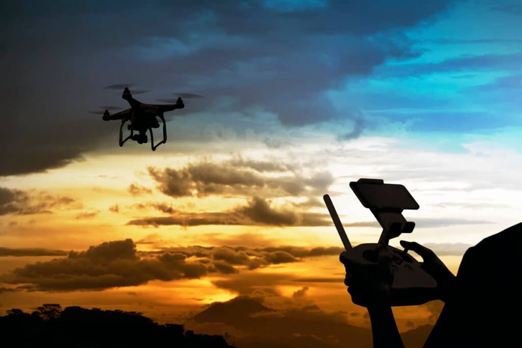 Silhouette d'un drone volant dans un ciel coloré au coucher du soleil, un pilote tenant la télécommande avec smartphone.