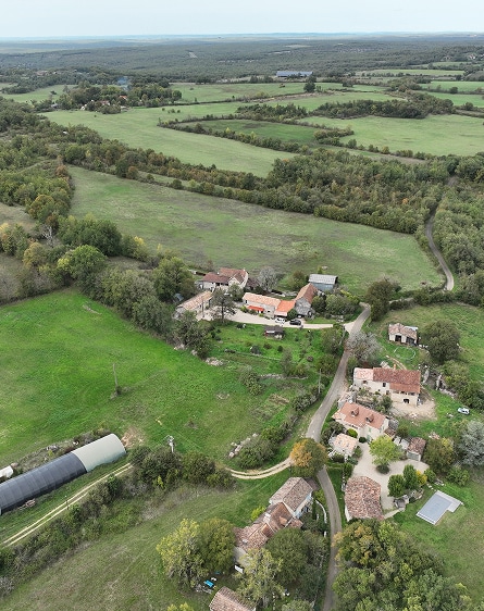 Vue aérienne d'un hameau dans la campagne, avec des maisons, une route sinueuse, des champs verdoyants et une serre.