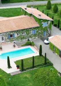 Vue aérienne d'une propriété ensoleillée : maisons couvertes de lierre, volets bleus, toits tuile, grande piscine, jardin verdoyant et voiture.