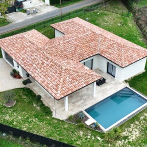 Maison contemporaine en L avec toit tuiles rouges, grande piscine bleue, terrasse carrelée et jardin luxuriant. Vue aérienne.
