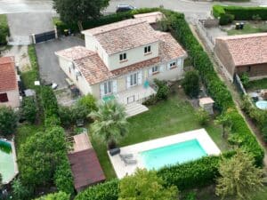 Vue aérienne d'une maison à toit de tuiles rouges avec piscine rectangulaire, terrasse carrelée, pelouse et haie dense dans un jardin.