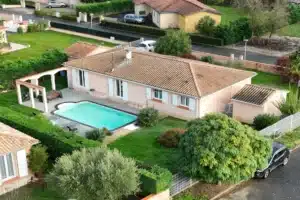 Vue aérienne d'une villa de plain-pied rose pâle avec une grande piscine rectangulaire et un jardin luxuriant, proche d'une rue résidentielle.