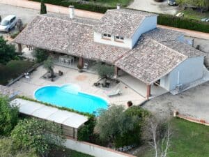 Vue aérienne d'une villa provençale avec toit en tuiles, piscine bleu vif, terrasse pavée, mobilier d'extérieur et arbres méditerranéens.