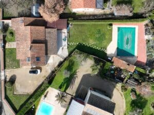 Vue aérienne d'une propriété résidentielle avec maison à toits de tuiles, deux piscines, jardin, arbres et voiture blanche garée.
