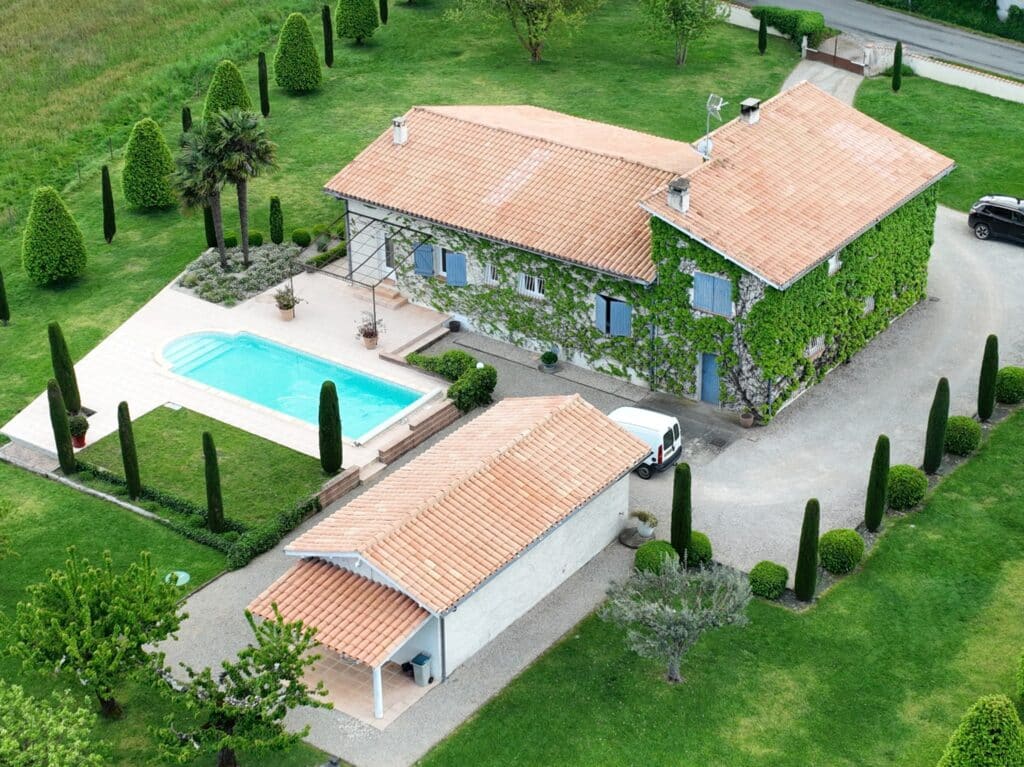 Maison de campagne avec piscine, façade en pierre couverte de lierre et jardin paysager avec cyprès.