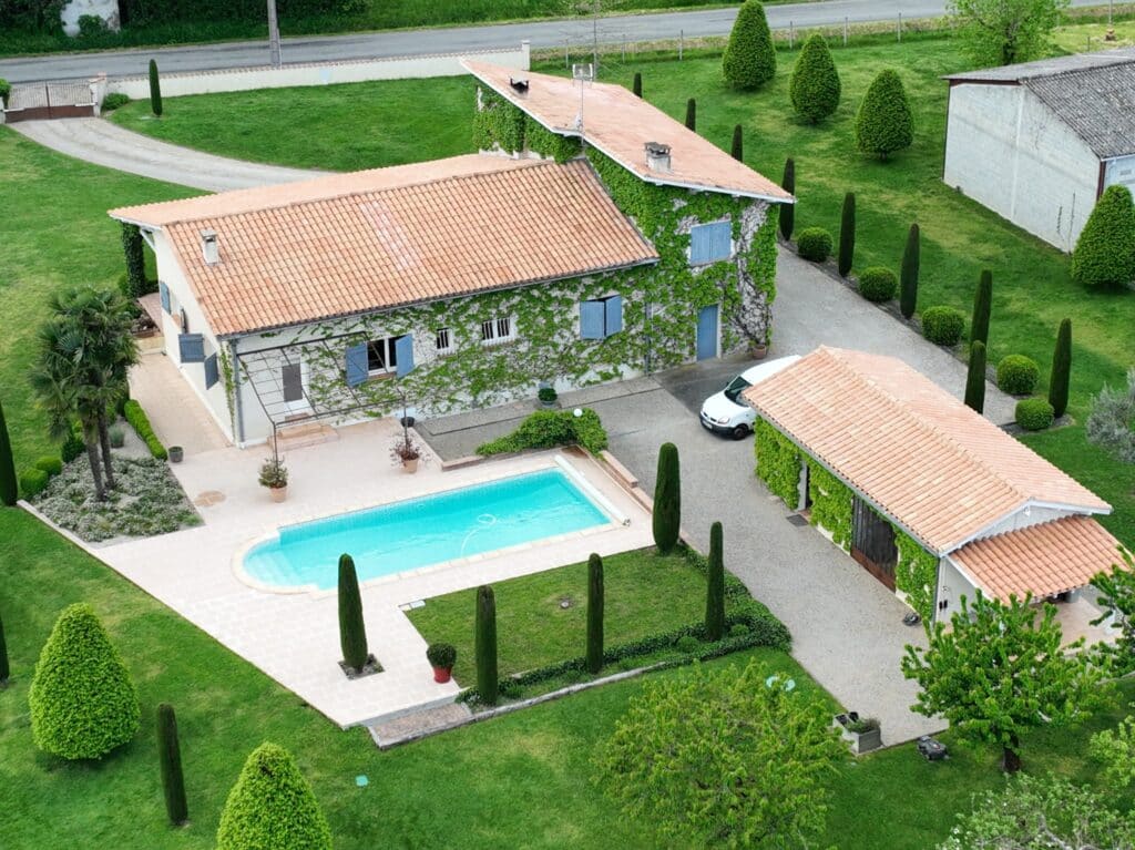 Grande maison avec piscine, murs couverts de lierre, volets bleus et jardin paysager avec cyprès au soleil.