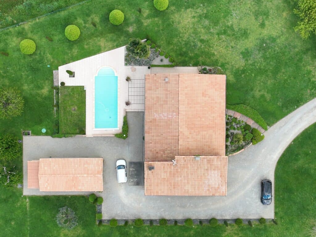Vue aérienne d'une villa avec piscine bleue, grand jardin verdoyant, toits en tuiles et voitures garées.