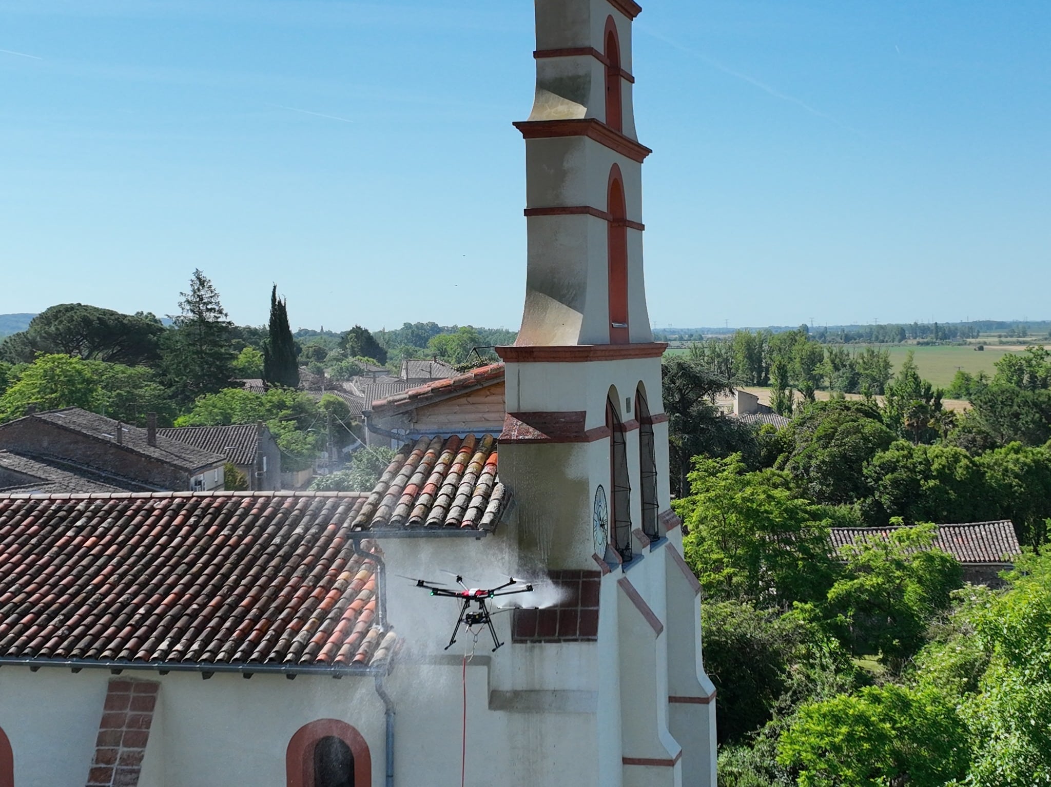 Drone pulvérisateur nettoyant la façade et le clocher d'une église ancienne sous un ciel bleu dégagé.