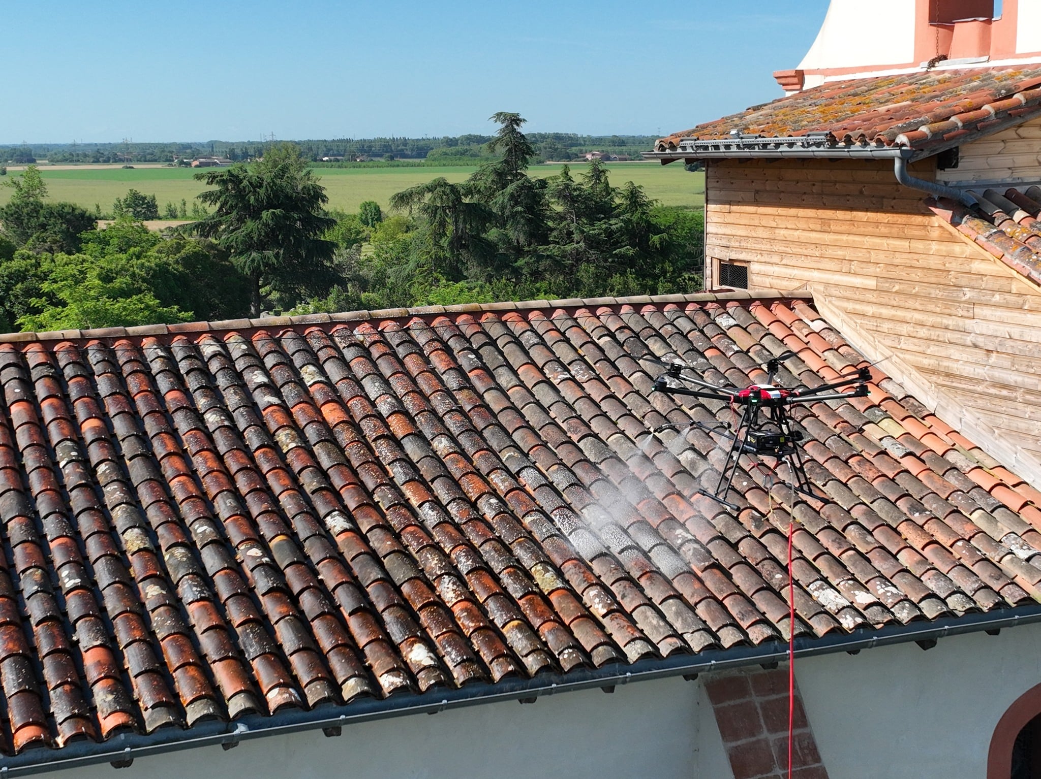 Drone de nettoyage pulvérisant un produit de démoussage sur une toiture en tuiles rouges en plein air.