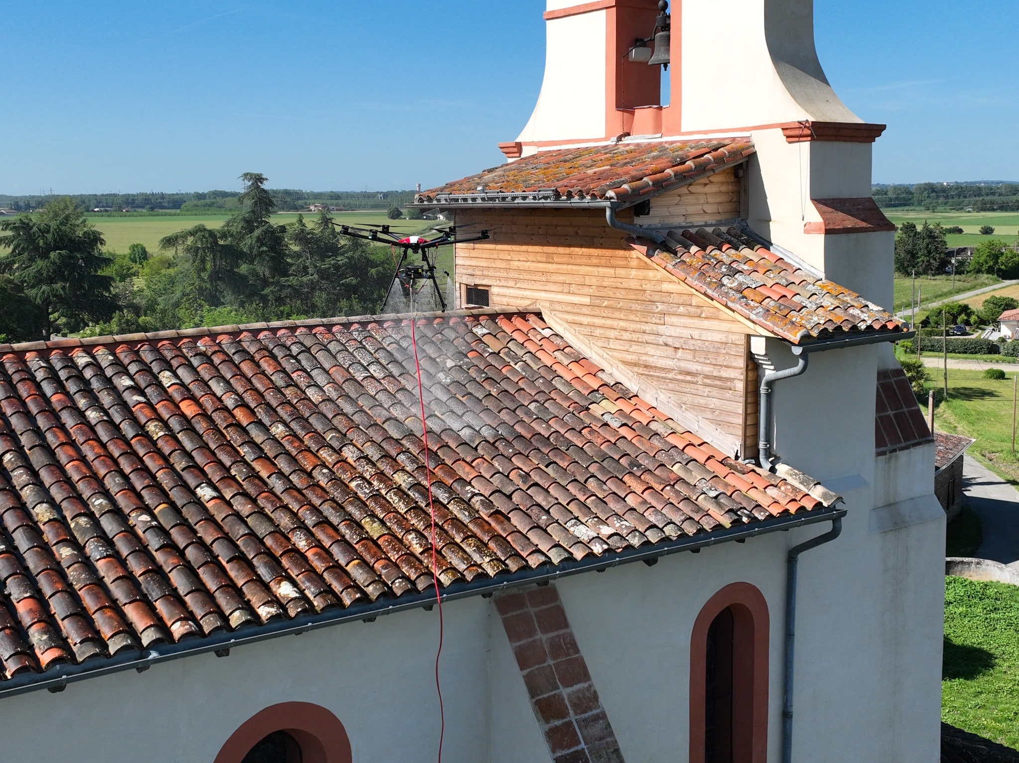 Drone de nettoyage pulvérisant un traitement sur les tuiles d'un clocher d'église par temps clair.